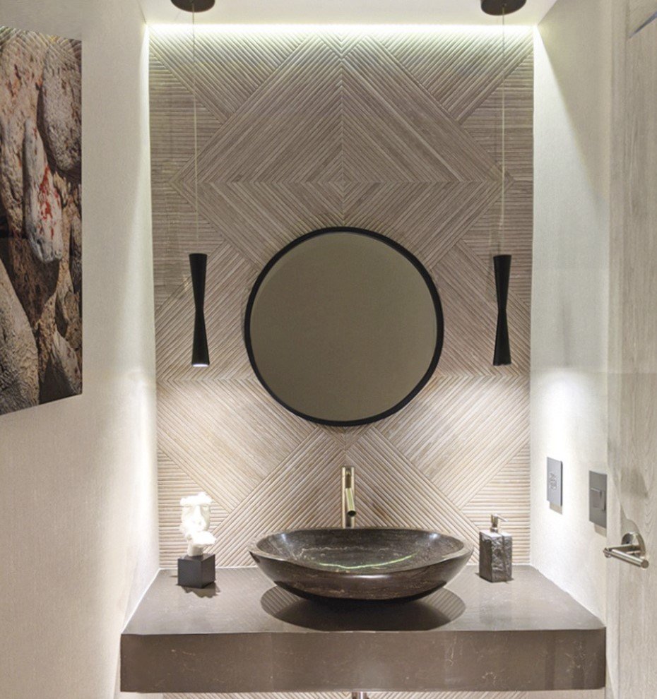 A backlit herringbone tile wall behind a stone vessel sink and round mirror